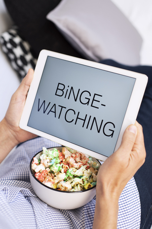 closeup of a young caucasian man lying comfortably on a couch, eating popcorn while is watching his tablet, with the text binge-watching in its screenの写真素材
