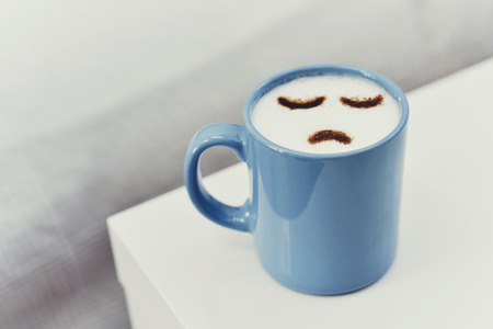 closeup of a blue cup of cappuccino with a sad face drawn with cocoa powder on its milk foam, on a white tableの写真素材