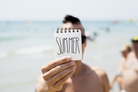 closeup of a young caucasian man in the beach showing a spiral notepad with the word summer handwritten in the first pageの写真素材