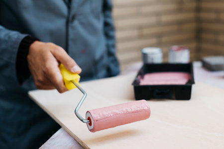 closeup of a young caucasian man, in a working coat, painting pink a wooden board with a paint...