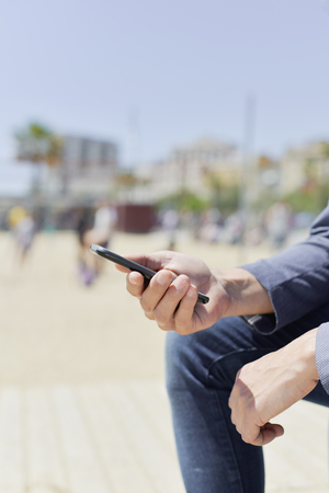 closeup of a young caucasian man, wearing a smart casual look, a pair of jeans and an elegant blue jacket, using a smartphone next to the beachの写真素材