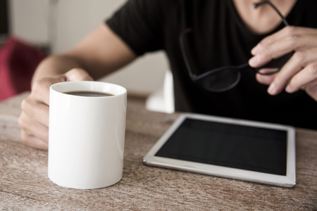 closeup of a young caucasian man, sitting at a table, using a tablet with a cup of coffee in one hand and a pair of eyeglasses in the other handの写真素材