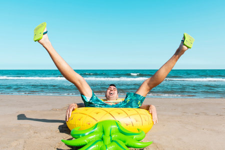 a laughing caucasian man sitting on a swim ring in the shape of a pineapple on the beach, with his...