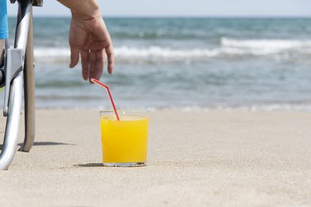 closeup of a young caucasian man relaxing on the beach, sitting on a deck chair next to a glass with a refreshing orange drink placed on the sand, with the sea in the backgroundの写真素材