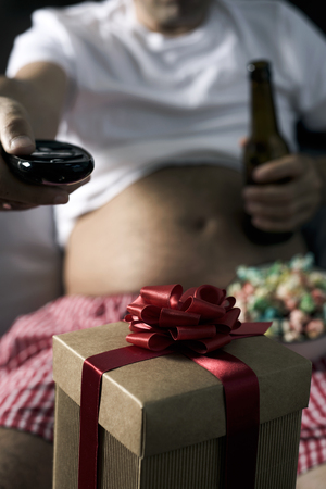 closeup of a gift and a caucasian man with a beer belly, sitting on the sofa, drinking beer and eating popcorn while is watching the televisionの写真素材