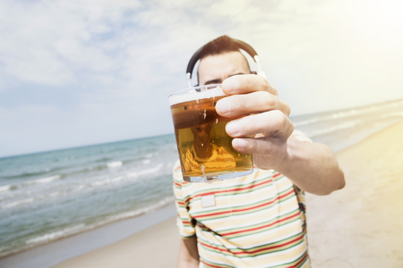 closeup of a young caucasian man on the beach listening to music with a pair of wireless headphones while is drinking a beer in front of the sea, with a sunbeam in the backgroundの写真素材