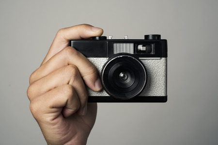 closeup of a caucasian man with a retro film camera in his hand, pointing to the obserever, about to press the shutter button, against an off-white backgroundの写真素材