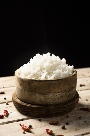 closeup of a boxwood bowl with some cooked long grain rice, on a rustic wooden table, against a black background, with some blank space on topの写真素材
