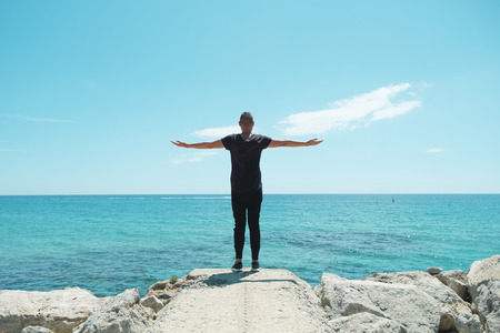 a young caucasian man, wearing black sweatpants and a black t-shirt, seen from behind with his arms in the air in front of the ocean, feeling freeの写真素材
