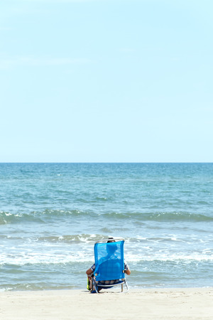 a caucasian man, seen from behind, relaxing on the beach, sitting on a deck chair and wearing a pair of green flip-flops, next to the water, with a blank space on the skyの写真素材