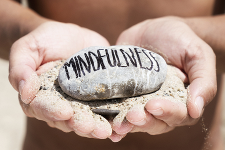 closeup of a young caucasian man, on the beach, with a handful of sand in his hands and a stone on top with the text mindfulness written in itの写真素材