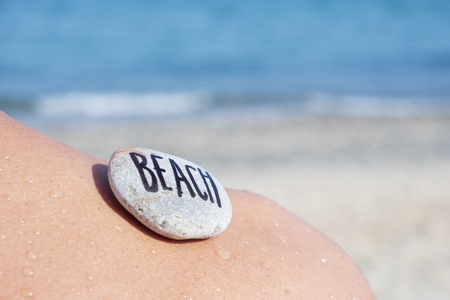 closeup of a young caucasian man seen from behind on the beach, in front of the ocean, with a stone on his back with the text beach written in itの写真素材