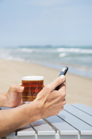 closeup of a young caucasian man, sitting at an outdoors table on the beach, using a smartphone while drinking a refreshing beerの写真素材
