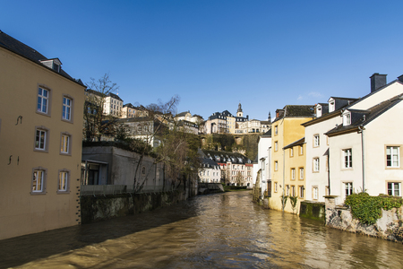 a view of the Alzette River as it passes through the Grund Quarter in Luxembourg City, Luxembourg, and the Ville Haute Quarter on the top, highlighting the belfry of the Saint-Michel churchの写真素材