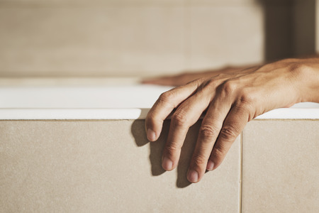 closeup of the hand of a young caucasian man on the side of a white bathtub while is taking a bath, in a beige bathroomの写真素材