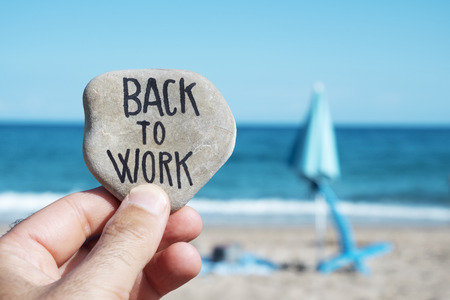 closeup of the hand of a young caucasian man on the beach, in front of a folded beach umbrella and the ocean in the background, holding a stone with the text back to work written in itの写真素材