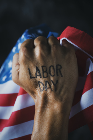 closeup of the raised fist of a young man with the text labor day handwritten in it, and the flag of the United Statesの写真素材