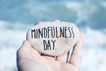 closeup of a young caucasian man, on the beach, showing a stone with the text mindfulness day written in itの写真素材
