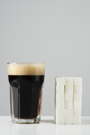 a glass of cola drink and a pile of white sugar cubes, disposed in a block, on an off-white background with some blank space on top, depicting the high sugar content of this kind of drinksの写真素材