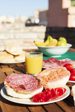 a sandwich with typical catalan pa amb tomaquet, bread with tomato, stuffed with salami, on a wooden table outdoors, next to a glass of orange juice and a bowl with fruitの写真素材
