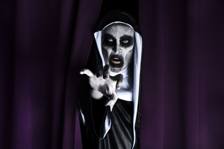 Closeup of a frightening evil nun, wearing a typical black and white habit, peeping out from a purple curtainの写真素材