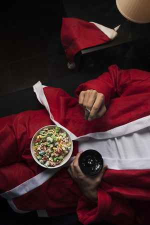 High angle view of a young man wearing a santa suit smoking a cigarette and eating popcorn in bedの写真素材