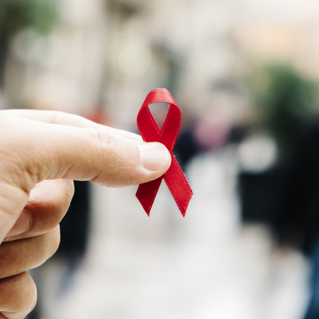 Closeup of a red awareness ribbon for the fight against AIDS in the hand of a young Caucasian man in a busy pedestrian street of a cityの写真素材