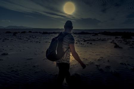 a young caucasian man seen from behind, carrying a backpack, walking by the sand of a desert, in backlit at duskの写真素材