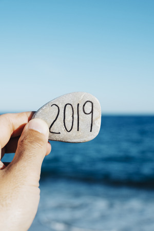closeup of a young man with a stone in his hand, with the number 2019, as the new year, handwritten in it, on the beach, with the ocean in the backgroundの写真素材