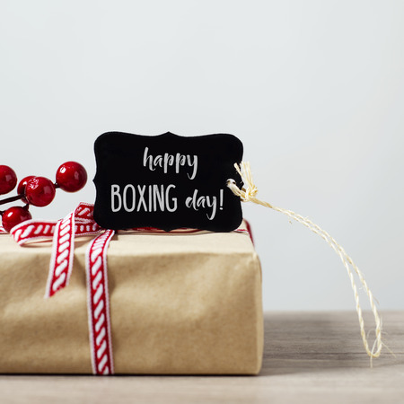 closeup of a cozy rustic gift, wrapped in kraft paper and tied with a red and white ribbon, ornamented with artificial red berries, and a black signboard with the text happy boxing day written in itの写真素材