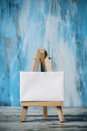 a blank white canvas on a wooden easel, on a wooden surface, against a blue rustic wooden backgroundの写真素材