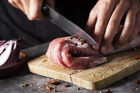 closeup of a young caucasian man cutting a piece of raw pork tenderloin with a knife, on a wooden chopping board, placed on a rustic wooden table or countertopの写真素材