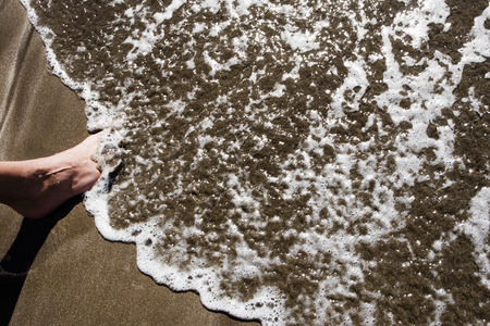 high angle view of the barefoot of a young caucasian man standing on the wet sand of the seashore covered by the foam of a waveの写真素材