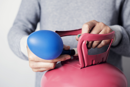 Closeup of a caucasian man, wearing a gray sweater, inflating a blue balloon with helium from a pink cylinderの写真素材