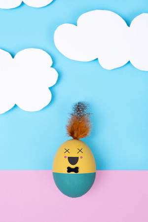 closeup of a handmade easter egg, with a cute face,  on a pink background, and a blue sky with white clouds in the backgroundの写真素材
