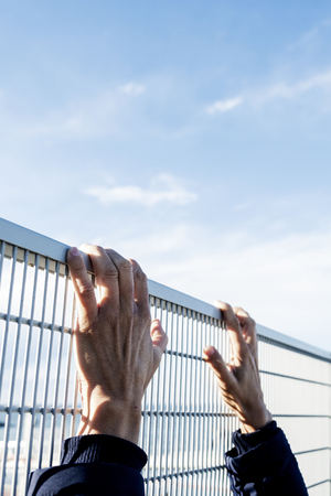 closeup of the hands of a man trying to climb up a metal fence, with the sky with a blank space on topの写真素材