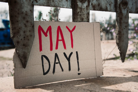 closeup of a brown cardboard signboard with the text may day handwritten in it, in the rusty fence...