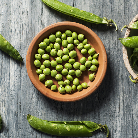 high angle view of some raw fresh green peas in an earthenware plate and some pea pods on a gray rustic wooden tableの写真素材