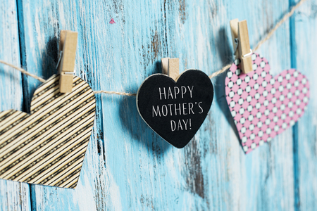 some hearts hung with clothespins in a hemp clothes line and a black heart-shaped signboard with the text happy mothers day written in it, against a blue rustic wooden backgroundの写真素材