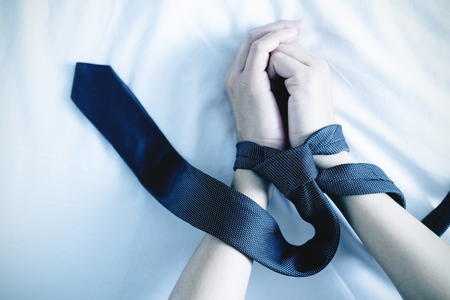 Closeup of the hands of a Caucasian man, lying down on a bed with white linen, tied up with a gray necktieの写真素材
