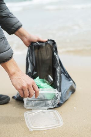 closeup of a caucasian man collecting garbage on a lonely beach, next to the water, as an action to clean the natural environmentの写真素材