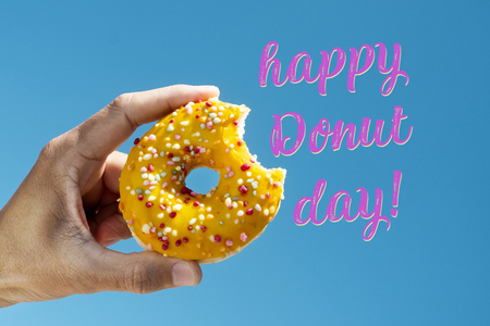 closeup of a nibbled appetizing donut, coated with a yellow frosting and sprinkles of different colors, in the hand of a young man and the text happy donut day against the blue skyの写真素材