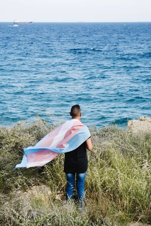 a young caucasian person, seen from behind, draping a transgender pride flag over his or her shoulders, facing the oceanの写真素材