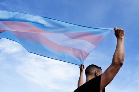 a young caucasian person, seen from behind, holding a transgender pride flag over his or her head against the blue skyの写真素材