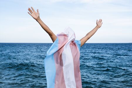a young caucasian person covered with a transgender pride flag with his or her arms open in the air, with the ocean in the backgroundの写真素材