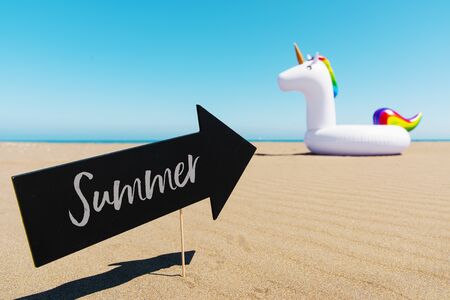 an arrow-shaped black signboard with the word summer written in it and a swim ring in the shape of a unicorn, on the sand of a beach, with the ocean in the backgroundの写真素材