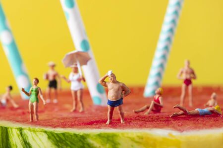 some miniature people wearing swimsuit relaxing on a refreshing ripe watermelon, with some white and blue drinking straws stuck in it, against a yellow backgroundの写真素材