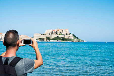 a young caucasian man, seen from behind, taking a picture of Calvi, in Corsica, France, with the...