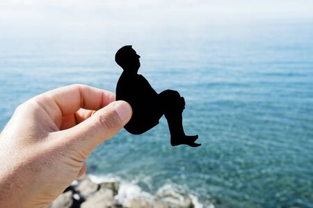 closeup of the hand of a man with a black paper cutout in the shape of a man cannonballing into the oceanの写真素材