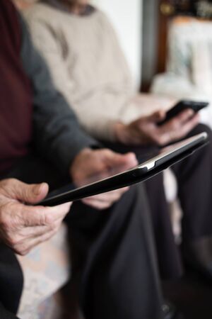 closeup of an old caucasian man using a tablet and an old caucasian man using a smartphone, sitting both in a couchの写真素材
